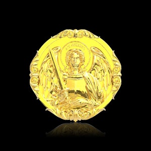 May include: A gold-coloured metal medallion with an ornate design featuring an angel with wings holding a sword. The angel is wearing a crown and has a halo.