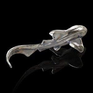 May include: A silver-colored sculpture of a shark with a long, curved tail. The shark is positioned on a black surface, with its reflection visible below.