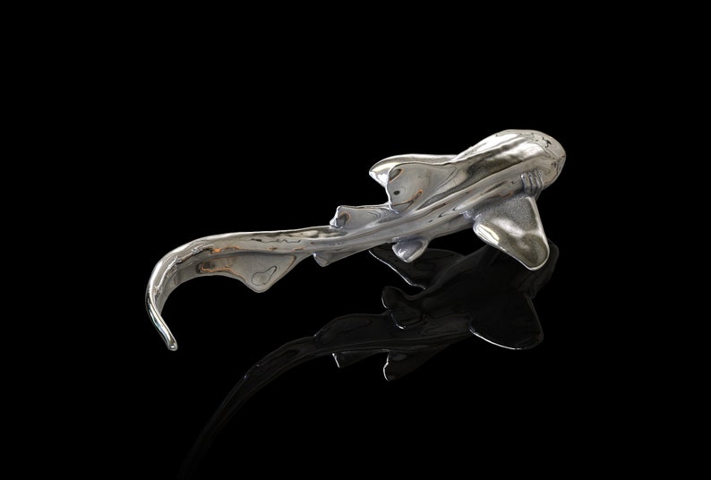 May include: A silver-colored sculpture of a shark with a long, curved tail. The shark is positioned on a black surface, with its reflection visible below.