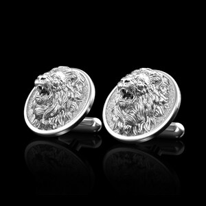 May include: A pair of silver cufflinks featuring a detailed lion head design. The lion's mouth is open and its mane is intricately sculpted.