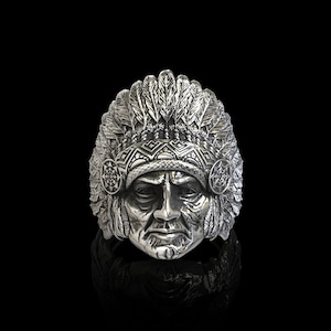 Puede incluir: Un anillo de plata con una representación detallada de un jefe nativo americano con un tocado. El anillo es una pieza llamativa con detalles intrincados y una representación realista del rostro del jefe.