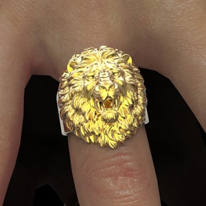 Puede incluir: Un anillo de cabeza de le&oacute;n de oro con un dise&ntilde;o detallado. La boca del le&oacute;n est&aacute; abierta y sus dientes son visibles. El anillo se lleva en un dedo.