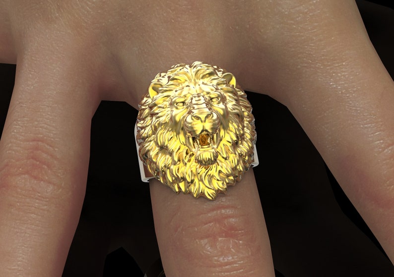 Puede incluir: Un anillo de cabeza de le&oacute;n de oro con un dise&ntilde;o detallado. La boca del le&oacute;n est&aacute; abierta y sus dientes son visibles. El anillo se lleva en un dedo.