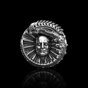 Puede incluir: Un anillo de plata con un diseño detallado de una cabeza humana rodeada por el cráneo de una criatura alienígena.