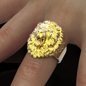 Puede incluir: Un anillo de oro con un dise&ntilde;o detallado de cabeza de le&oacute;n. La boca del le&oacute;n est&aacute; abierta, mostrando sus dientes. El anillo se lleva en un dedo.