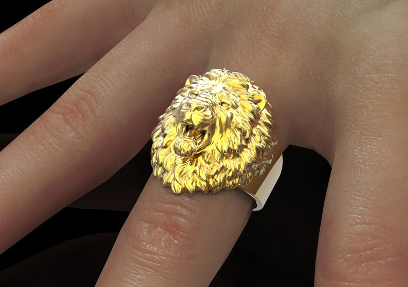 Puede incluir: Un anillo de oro con un dise&ntilde;o detallado de cabeza de le&oacute;n. La boca del le&oacute;n est&aacute; abierta, mostrando sus dientes. El anillo se lleva en un dedo.