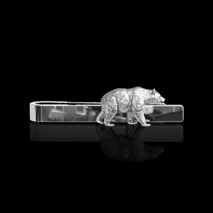 Puede incluir: Pinza de corbata de plata con una figura de oso detallada. El oso camina hacia la derecha y tiene una superficie texturizada.