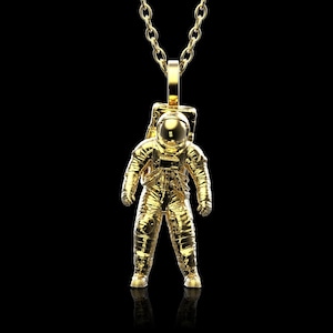 Può includere: Una collana con un ciondolo a forma di astronauta in oro. L'astronauta indossa una tuta spaziale e un casco. La collana è su una catena d'oro.