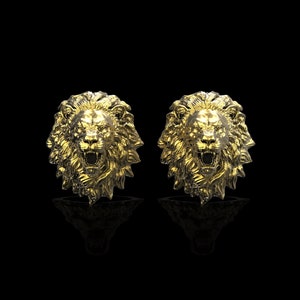 May include: A pair of gold lion head earrings. The lion heads are facing forward with their mouths open and their teeth showing.