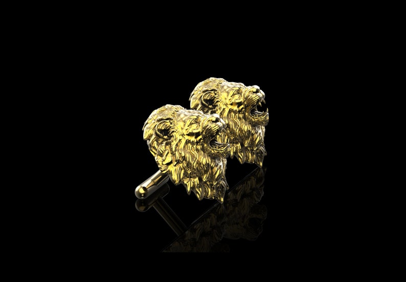May include: A pair of gold lion head cufflinks. The lion heads are facing forward with their mouths open, as if roaring. The cufflinks are made of a shiny metal and have a detailed design.