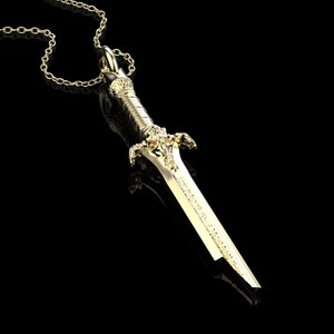May include: A gold-coloured sword pendant necklace with a detailed design. The sword has a pointed tip and a decorative handle.