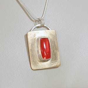 May include: A rectangular pendant necklace featuring a polished, vibrant red stone set in a brushed gold-tone metal plate. The pendant is suspended from a silver-tone snake chain by a semi-circular metal bail, creating a clean, structured look.