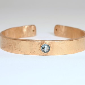 May include: A hammered copper cuff bracelet with a light blue gemstone set in the center. The open-ended bracelet has a textured surface and a warm, metallic color. The gemstone is round and appears to be a light blue color.
