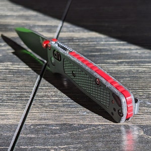 Puede incluir: Un cuchillo de bolsillo negro con un acento rojo a lo largo del borde del mango. El cuchillo tiene un mango texturizado y una hoja parcialmente abierta.