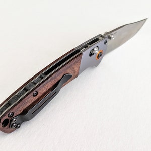 May include: A brown wood handled pocket knife with a silver blade and a black pocket clip. The knife is closed and the blade is visible.