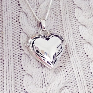 Puede incluir: Un colgante plateado en forma de corazón sobre una cadena fina. El relicario está muy pulido y refleja la luz, con un diseño intrincado alrededor de los bordes. El colgante cuelga sobre un suéter de punto de cable gris claro.
