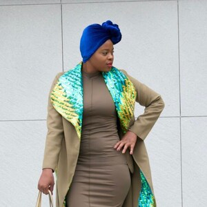 May include: A woman wearing a brown coat with a green and gold sequined lining. She is also wearing a blue turban, a brown dress, and gold shoes. She is carrying a gold handbag with a green and gold sequined design.