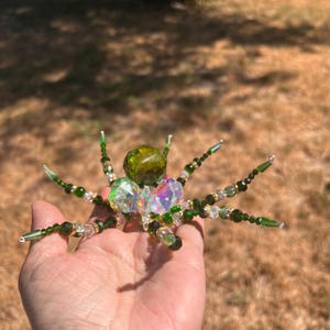 May include: A decorative spider ornament crafted from faceted green and iridescent clear beads. The body features a large dark green central bead, while the eight legs are constructed from smaller green and clear beads on wire.