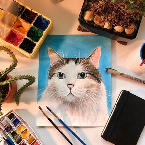 K&ouml;nnte beinhalten: Ein Gem&auml;lde einer wei&szlig;en und braunen Katze mit blauen Augen. Die Katze ist auf einem blauen Hintergrund gemalt. Das Gem&auml;lde befindet sich auf einem Tisch mit Kunstmaterialien, darunter Pinsel, eine Aquarellpalette und ein schwarzes Notizbuch.