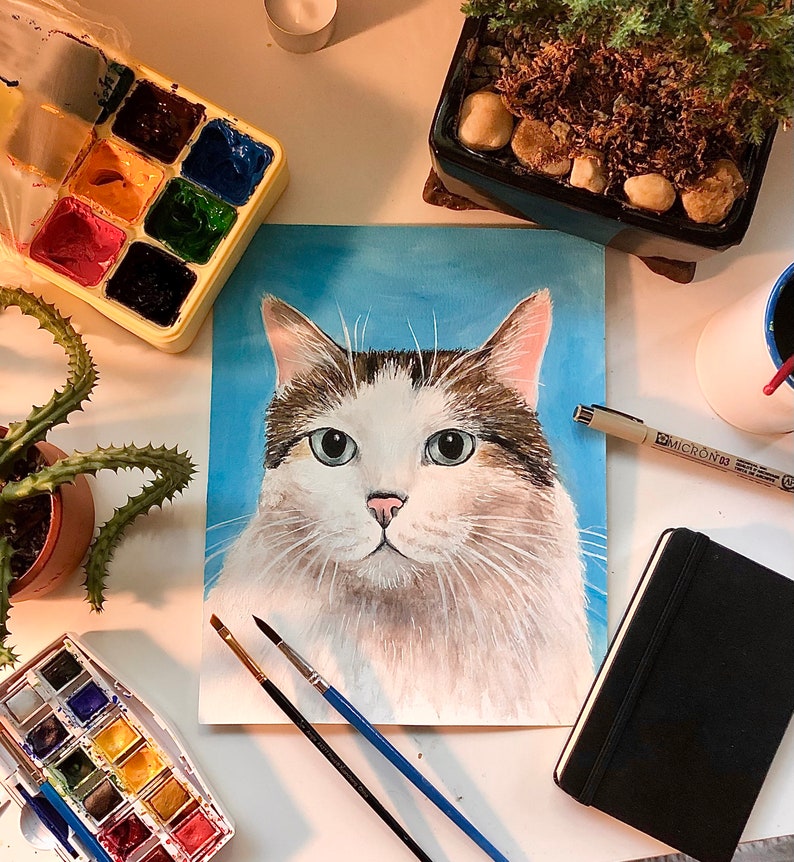 K&ouml;nnte beinhalten: Ein Gem&auml;lde einer wei&szlig;en und braunen Katze mit blauen Augen. Die Katze ist auf einem blauen Hintergrund gemalt. Das Gem&auml;lde befindet sich auf einem Tisch mit Kunstmaterialien, darunter Pinsel, eine Aquarellpalette und ein schwarzes Notizbuch.