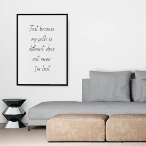 May include: A framed black and white inspirational quote on a white wall. The quote reads "Just because my path is different, does not mean I'm lost". A grey sofa with two tan footstools sits in front of the quote.