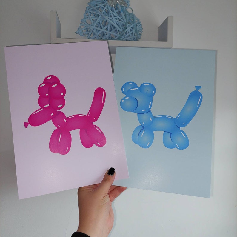 May include: Two prints featuring balloon animal illustrations. One print has a pink poodle-shaped balloon on a light pink background. The other print has a blue dog-shaped balloon on a light blue background. The prints are held by a hand.