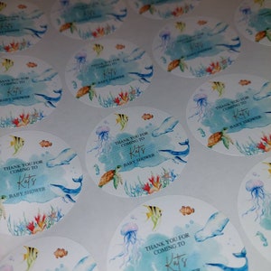 May include: Round blue and white thank you stickers with a watercolor design of sea life, including a whale, jellyfish, and a turtle. The text on the stickers reads "Thank you for coming to Kats Baby Shower".