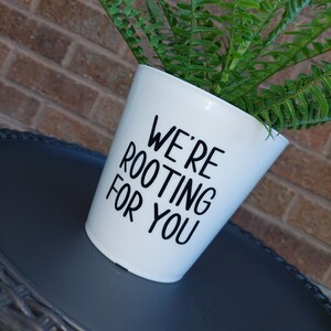 May include: White ceramic planter with black text that reads "We're rooting for you". A green fern plant is growing in the planter.