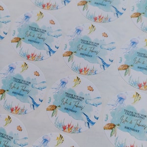 May include: Round, blue and white stickers with an ocean theme. The stickers feature a watercolor design of sea life, including a turtle, jellyfish, and fish. The text on the stickers reads "Thank you for celebrating Isla & Tobias' Birthday."