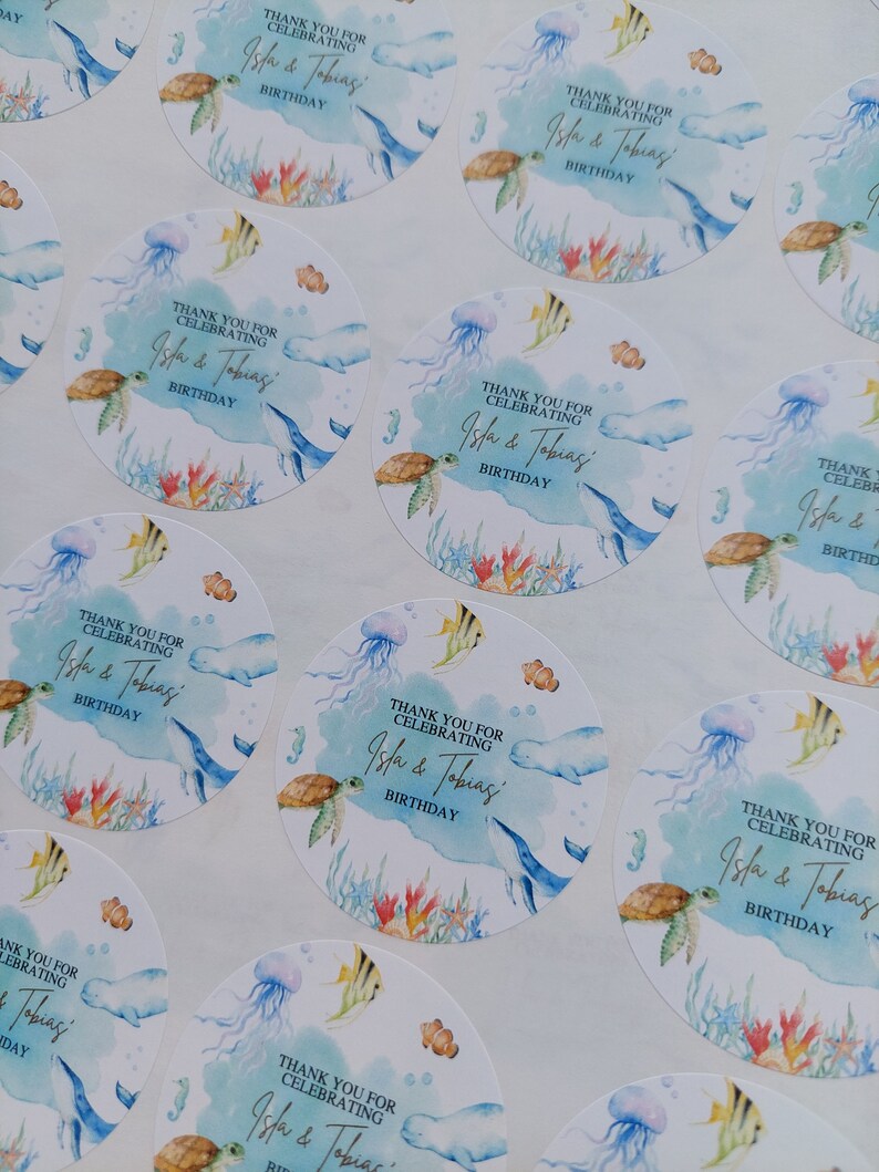 May include: Round, blue and white stickers with an ocean theme. The stickers feature a watercolor design of sea life, including a turtle, jellyfish, and fish. The text on the stickers reads "Thank you for celebrating Isla & Tobias' Birthday."