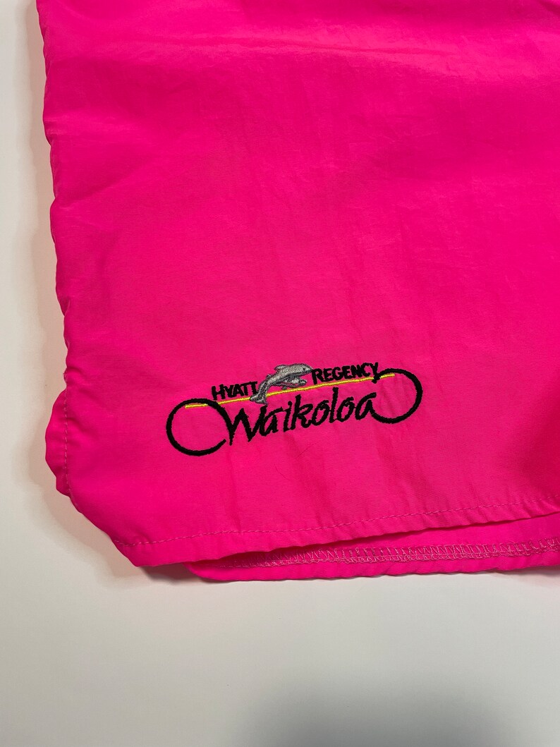 May include: Pink swim trunks with an embroidered logo that says "Hyatt Regency Waikoloa" with a dolphin graphic.