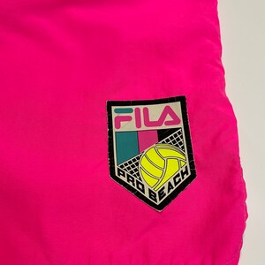 May include: Pink swim trunks with a black Fila Pro Beach logo featuring a volleyball. The logo has a white volleyball and a blue, pink, and green background.