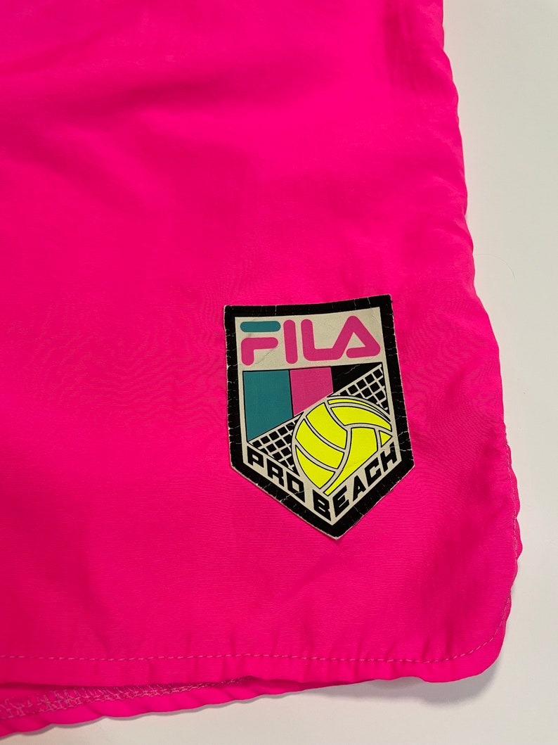 May include: Pink swim trunks with a black Fila Pro Beach logo featuring a volleyball. The logo has a white volleyball and a blue, pink, and green background.
