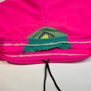 May include: Pink swim trunks with a green and white waistband and a logo that says "Pro Beach" in black and yellow lettering.