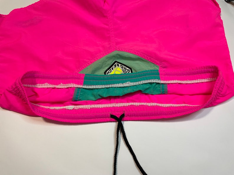 May include: Pink swim trunks with a green and white waistband and a logo that says "Pro Beach" in black and yellow lettering.