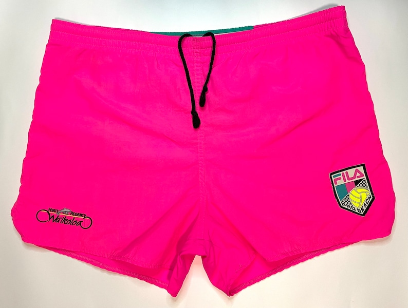 May include: Pink swim trunks with a black drawstring waist and a Fila logo on the right leg. The logo is a shield with a volleyball and the word "Fila" in white letters. The left leg has a small embroidered logo that says "Hotel Regency Waikoloa".