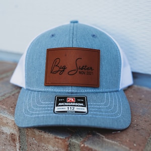 May include: A blue and white trucker hat with a brown leather patch that says "Big Sister Nov. 2021".