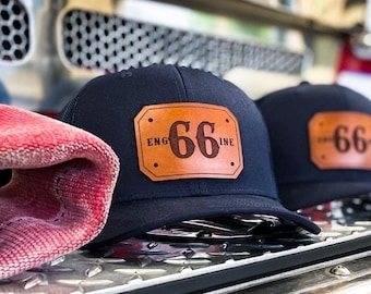 May include: A navy blue baseball cap featuring a rectangular tan leather patch on the front. The patch is embossed with the text "ENGINE 66" in a dark brown font. The cap is displayed alongside other identical caps on a metallic surface.