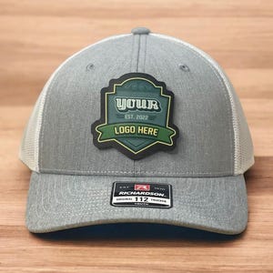 May include: A heather gray trucker hat with a white mesh back and a dark green shield-shaped patch on the front. The patch features the text YOUR, EST. 2022, and LOGO HERE. A black sticker on the brim reads RICHARDSON 112 TRUCKER YOUTH.
