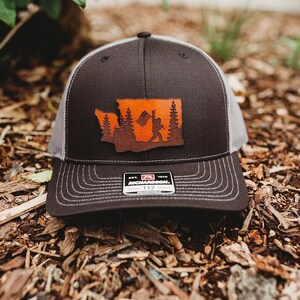 May include: A black and gray trucker hat with a brown leather patch featuring an outline of the state of Washington with a silhouette of a person holding a flag inside the state outline. The patch has a brown background with orange accents. The hat has a black mesh back and a white logo that says "Richardson 112".
