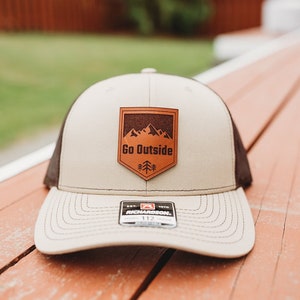 May include: A beige and brown trucker hat with a brown leather patch that says "Go Outside" and features a mountain silhouette. The patch is in the shape of a shield.