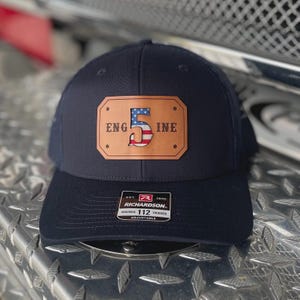 K&ouml;nnte beinhalten: Eine marineblaue Trucker-Cap mit einem rechteckigen, hellbraunen Lederaufn&auml;her auf der Vorderseite. Der Aufn&auml;her zeigt den Text "ENG" und "INE" neben einer gro&szlig;en "5", die mit einem US-Flaggenmuster gef&uuml;llt ist. Ein schwarz-wei&szlig;es Richardson 112 Etikett ist angebracht.
