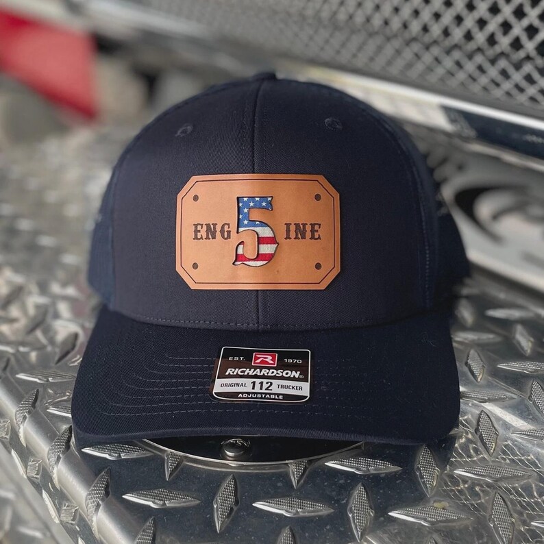 K&ouml;nnte beinhalten: Eine marineblaue Trucker-Cap mit einem rechteckigen, hellbraunen Lederaufn&auml;her auf der Vorderseite. Der Aufn&auml;her zeigt den Text "ENG" und "INE" neben einer gro&szlig;en "5", die mit einem US-Flaggenmuster gef&uuml;llt ist. Ein schwarz-wei&szlig;es Richardson 112 Etikett ist angebracht.