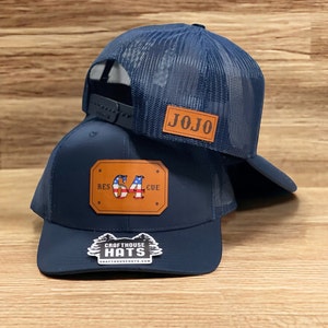 May include: Two navy blue trucker hats with leather patches. The top hat has a brown leather patch with the name "JOJO" in white lettering. The bottom hat has a brown leather patch with the number "64" in white lettering and the words "RESCUE" and "CUE" in white lettering on either side of the number. The bottom hat also has a small sticker with the words "CRAFTHOUSE HATS" and a logo of a pine tree.