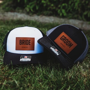 May include: Two black and white trucker hats with brown leather patches. The patch on the white hat says "BRIDE" with the date "09.08.2013". The patch on the black hat says "GROOM" with the same date.