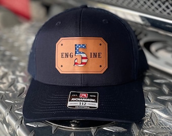 May include: A navy blue trucker hat with a brown leather patch that says "ENG 5 INE" with a small American flag in the number 5. The hat has a black mesh back and a black adjustable strap with a Richardson logo and the text "EST. 1970 ORIGINAL 112 TRUCKER ADJUSTABLE".