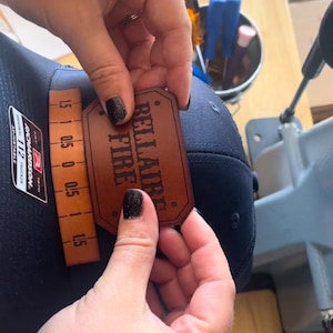 May include: A brown leather patch with the text "RELLAIRE FIRE" in a dark brown font. The patch is being held by a hand.