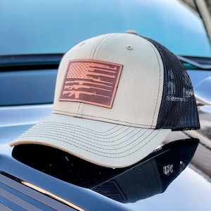 May include: A beige and black trucker hat with a brown leather patch featuring a stylized American flag with guns in place of the stripes.
