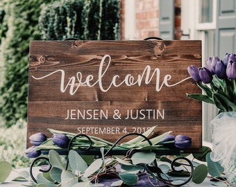 May include: A wooden welcome sign with the text "Welcome Jensen & Justin September 4, 2019" in white lettering. The sign is on a table with purple flowers and greenery.