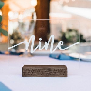 May include: A clear acrylic table number with the word "nine" written in white calligraphy on a wooden base.